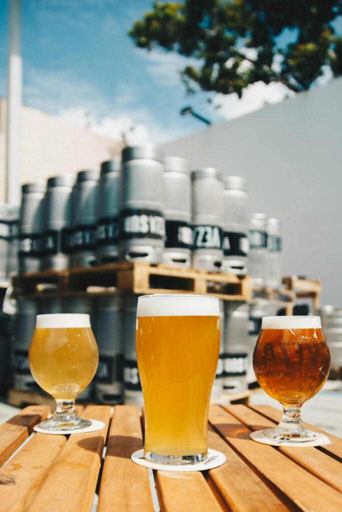 Assorted beer glasses on a wooden table with brewery kegs in the background.